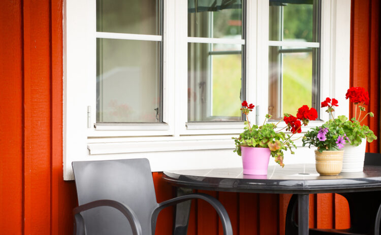 Bild på fönstret på en röd stuga, väggen är röd och framför fönstret står ett bord med blommor på.