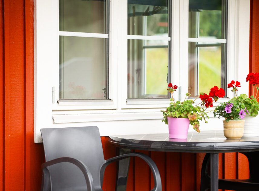 Bild på fönstret på en röd stuga, väggen är röd och framför fönstret står ett bord med blommor på.