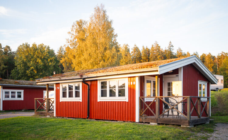 Bild på röd stuga sedd snett framifrån, i bakgrunden skymtas fler stugor och träd mot en blå himmel.