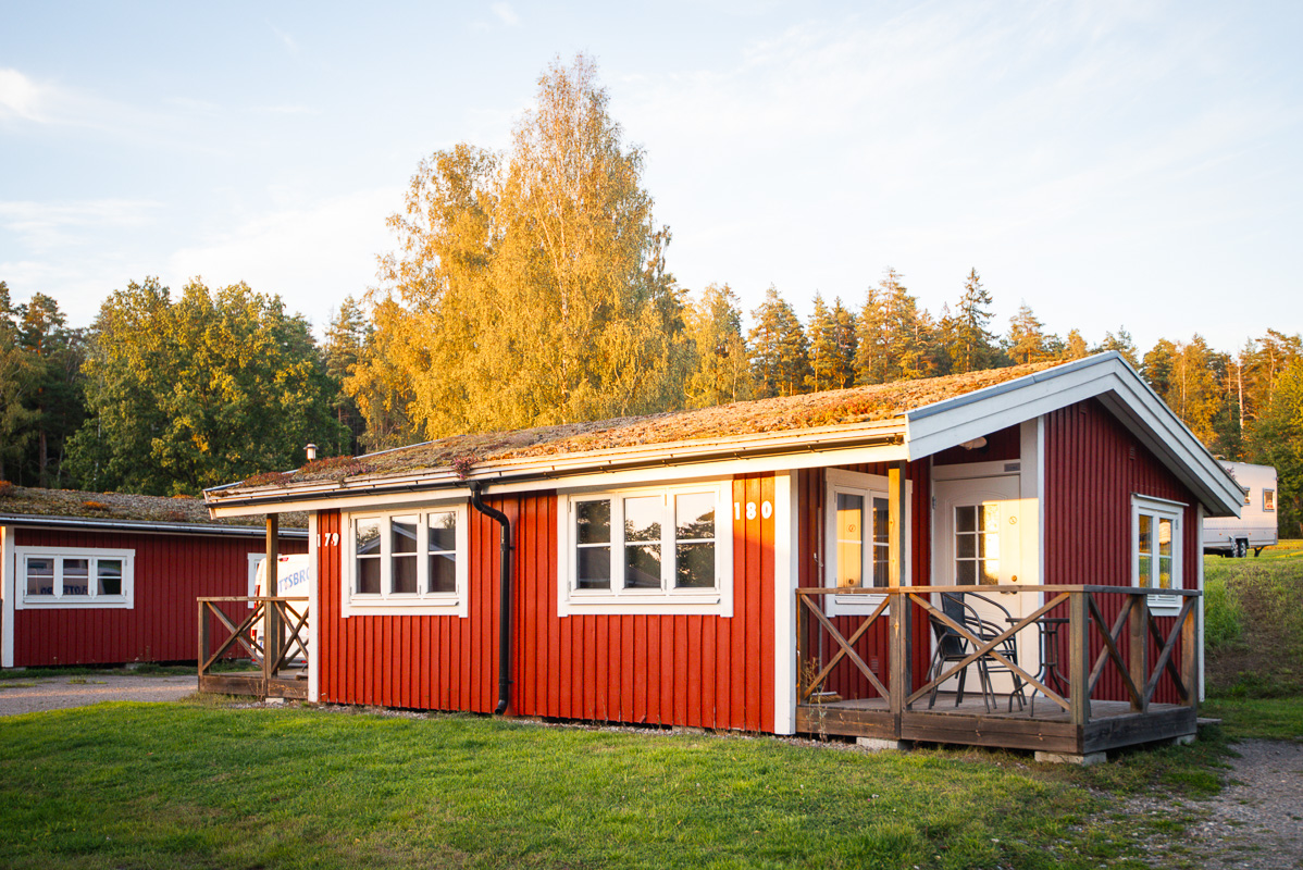 Bild på röd stuga sedd snett framifrån, i bakgrunden skymtas fler stugor och träd mot en blå himmel.