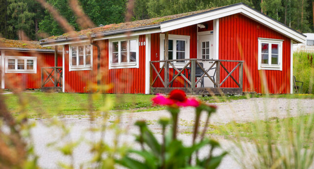 Röd stuga sedd snett framifrån, i förgrunden syns en rosa blomma och bakom stugan syns gröna träd.