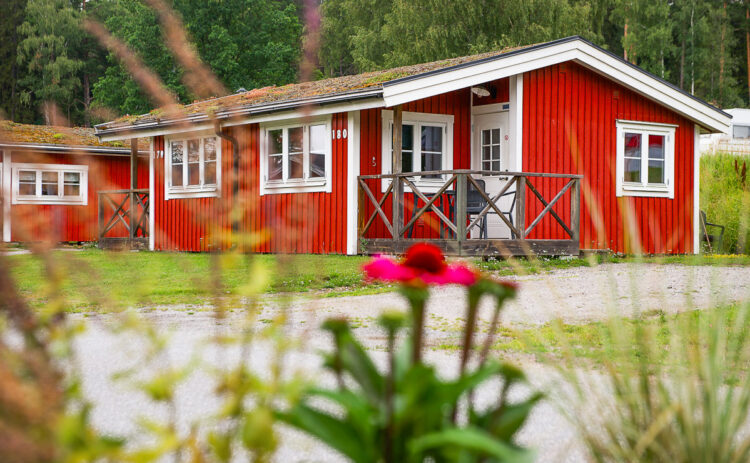 Röd stuga sedd snett framifrån, i förgrunden syns en rosa blomma och bakom stugan syns gröna träd.