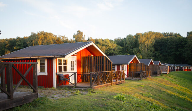 Bilden visar röda stugor i solnedgången i bakgrunden syns en grön skog, himlen är blå.