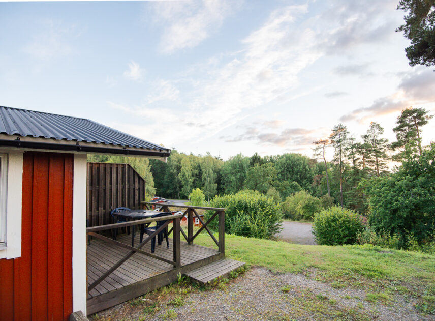 Röd stuga sedd från sidan med utsikt över en slänt och kulle i bakgrunden, det är sommar och grönska, himlen är blå.
