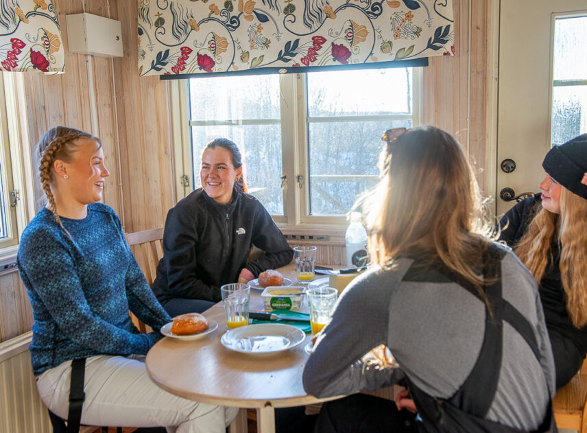 Fyra tjejer sitter kring ett bord i en liten stuga, det kommer in solljus genom ljusa fönster.