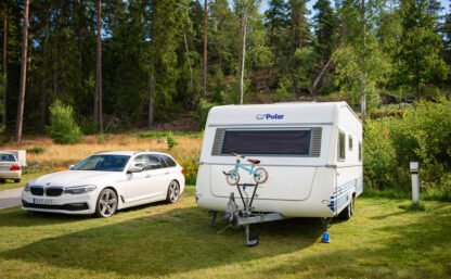 Bild på husvagn och bil parkerade bredvid varandra på en grön gräsmatta, i bakgrunden syns barrskog och himlen skymtar mellan träden.