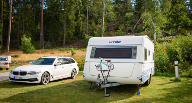 Bild på husvagn och bil parkerade bredvid varandra på en grön gräsmatta, i bakgrunden syns barrskog och himlen skymtar mellan träden.