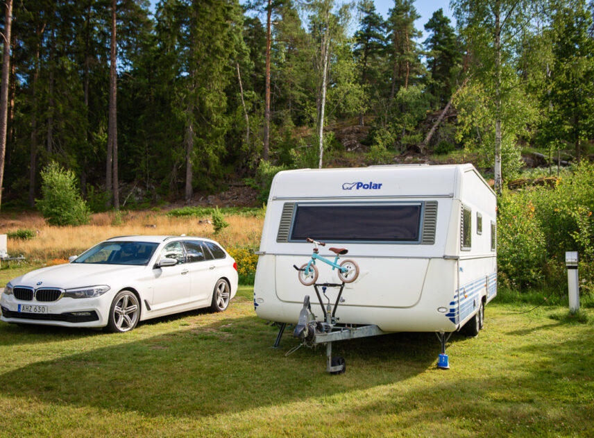 Bild på husvagn och bil parkerade bredvid varandra på en grön gräsmatta, i bakgrunden syns barrskog och himlen skymtar mellan träden.