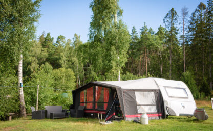 Husvagn med förtält parkerade på en grön gräsmatta omgiven av skog och blå himmel.