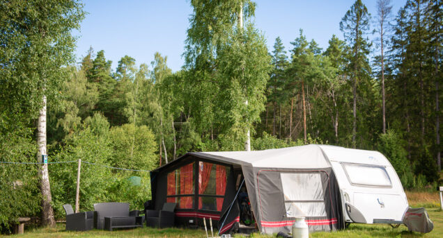 Husvagn med förtält parkerade på en grön gräsmatta omgiven av skog och blå himmel.