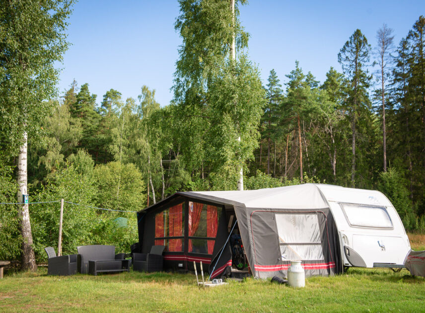 Husvagn med förtält parkerade på en grön gräsmatta omgiven av skog och blå himmel.