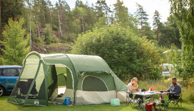 Bild på en familj som sm sitter vid ett bord utanför ett grönt tält på sommaren, i bakgrunden syns grönskande buskar och en skog.