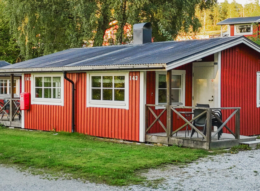 Röd stuga sedd snett framifrån bakom skymtas 2 andra liknande stugor omgivna av grönska.
