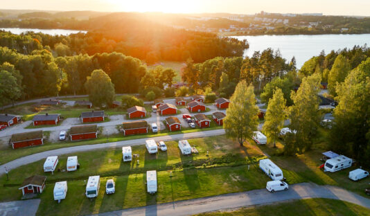 Camping Stockholm vid sjö och natur–Vy av campingplats med husvagnar och stugor i solnedgången omgiven av skog, i bakgrunden syns en sjö.