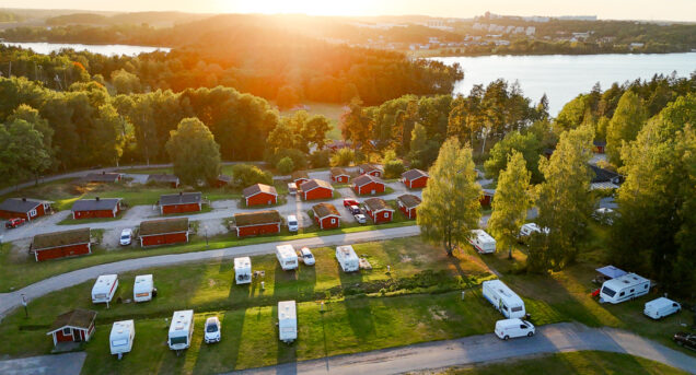 Camping Stockholm vid sjö och natur–Vy av campingplats med husvagnar och stugor i solnedgången omgiven av skog, i bakgrunden syns en sjö.