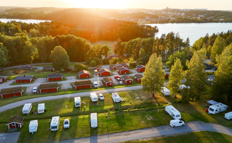 Camping Stockholm vid sjö och natur–Vy av campingplats med husvagnar och stugor i solnedgången omgiven av skog, i bakgrunden syns en sjö.
