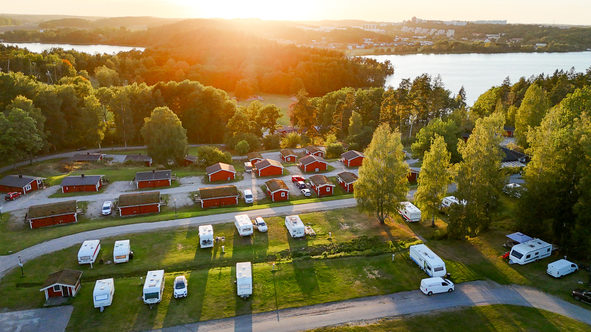 Vy av campingplats med husvagnar och stugor i solnedgången omgiven av skog, i bakgrunden syns en sjö.