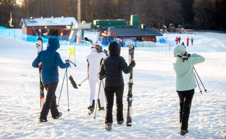 Fyra personer sedda bakifrån som bär på skidutrustning, de promenerar mot liften i en slalombacke.
