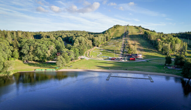 bilden visar Flottsbros backe på sommaren, ett grönskande landskap med blått vatten och klarblå himmel.