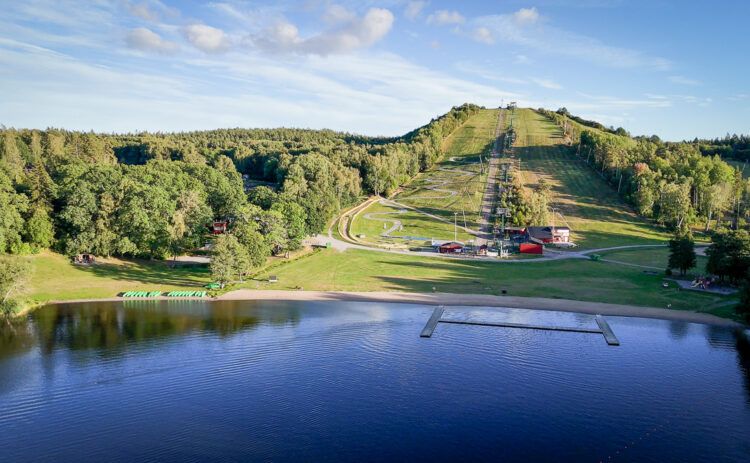 bilden visar Flottsbros backe på sommaren, ett grönskande landskap med blått vatten och klarblå himmel.