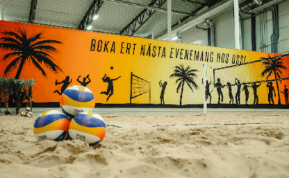 Bilden visar en beachvolleybana inomhus med tre bollar och en orange skylt i bakgrunden.