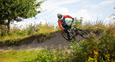 Cyklist som cyklar i Bike Park, i förgrunden syns grönskande blommor och himlen är blå.