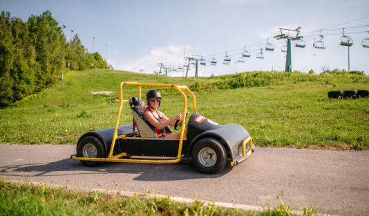 Bilden visar Flottsbro Downhill Cars, aktivitet med gokart sedd från sidan kör på en väg i en backe med grönt gräs och blå himmel bakom.