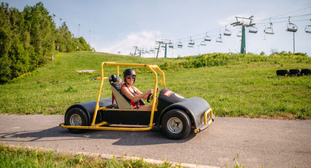 Bilden visar Flottsbro Downhill Cars, aktivitet med gokart sedd från sidan kör på en väg i en backe med grönt gräs och blå himmel bakom.
