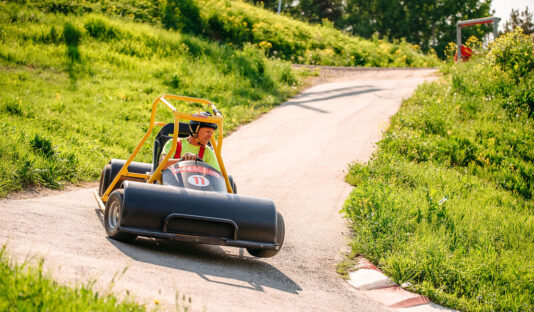 En gokart med förare som kör nedför en bilväg i en backe en grönskande sommardag med blå himmel.
