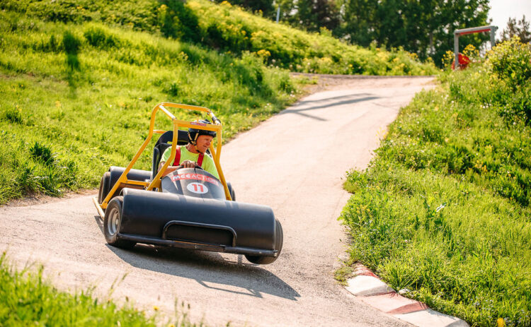 En gokart med förare som kör nedför en bilväg i en backe en grönskande sommardag med blå himmel.