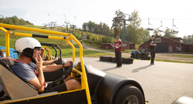 Bild med en gokart och förare från sidan frmför och längre bort i bild står en person som ger instruktioner.