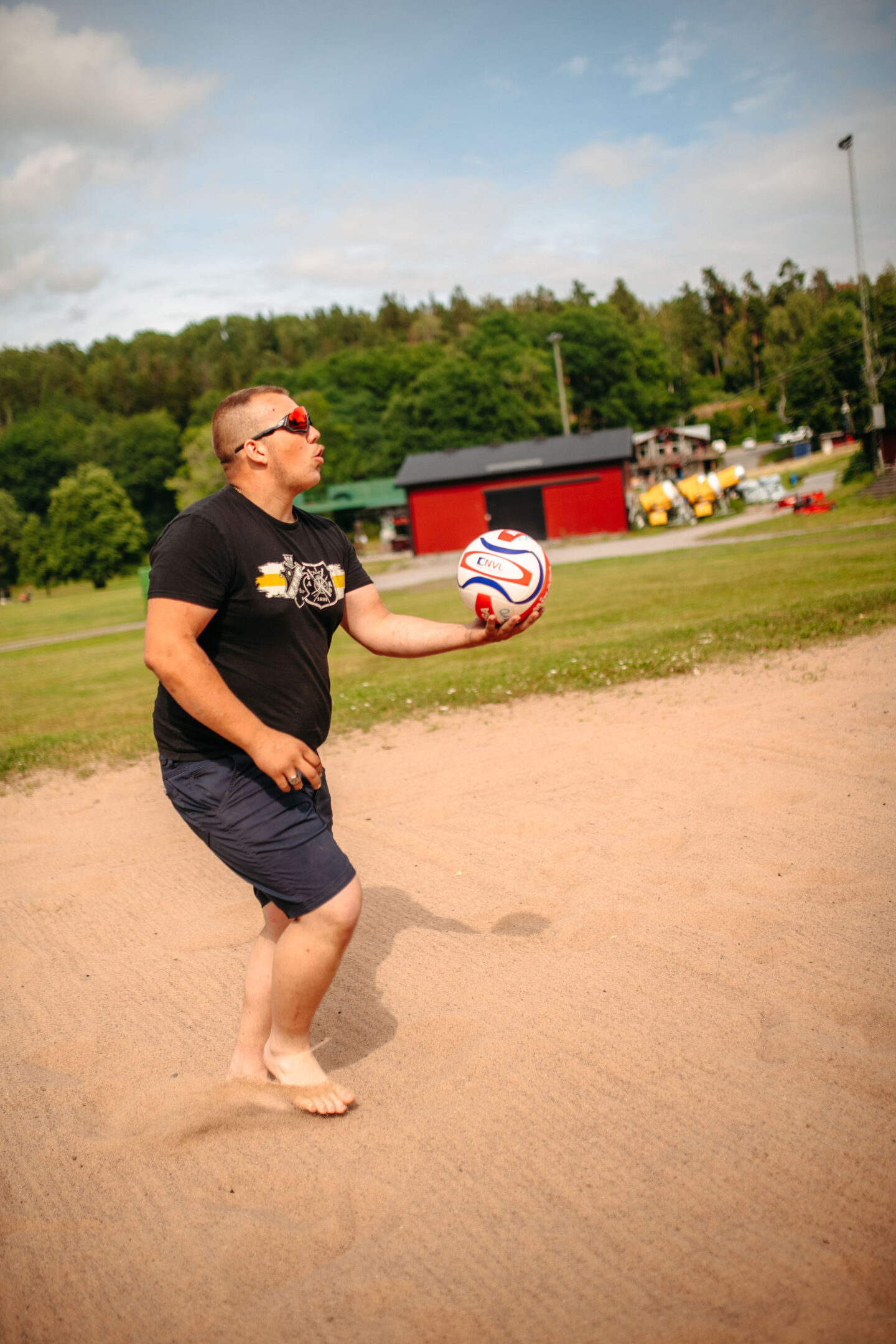 En person rörelse som håller en beachvolleyboll, i backgrunden syns grönska och en röd byggnad.