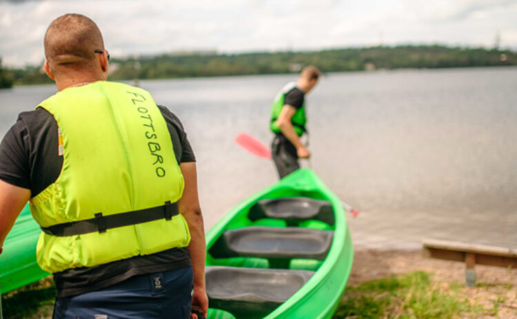 Två män sedda bakifrån bär en kanot ner till en sjö en sommardag