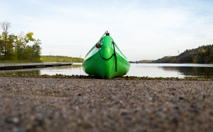 A canoe laying on shore of a beach, in the background you see a lake and nature.