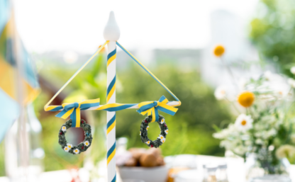 Bolden visar en svensk midsommarstång i miniatyr på ett dukat bord med blommor mot grönskande ljus bakgrund.