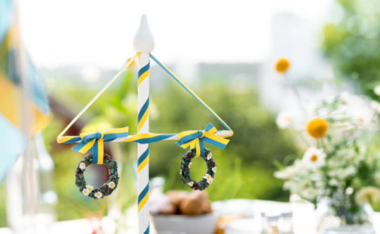 Bolden visar en svensk midsommarstång i miniatyr på ett dukat bord med blommor mot grönskande ljus bakgrund.