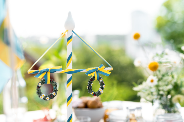 Bilden visar en svensk midsommarstång i miniatyr på ett dukat bord med blommor mot grönskande ljus bakgrund.