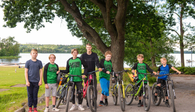 Porträtt på en grupp ungdomar och en ledare i mitten, några håller i cyklar och de står under en grönskande ek.