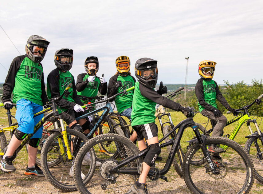 Bild på en grupp ungdomar på cyklar som är klädda i full downhill utrustning, ett grönskande landskap med blå himmel i bakgrunden.