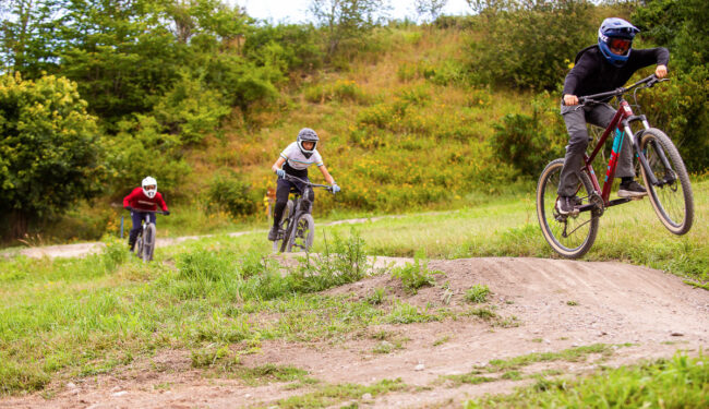 Tre cyklister som cyklar över hopp, i bakgrunden syns buskage och grönskande gräs.
