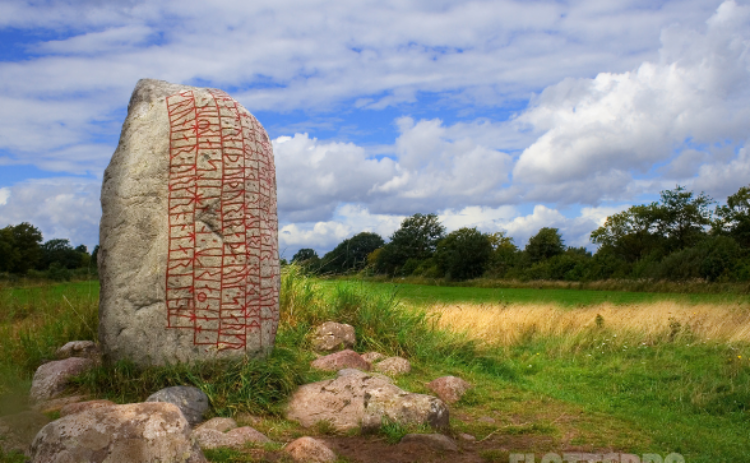 Bild på runsten i ett grönskande landskap med blå himmel