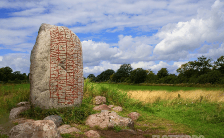 Bild på runsten mot bakgrund av grön äng, träd och blå himmel.