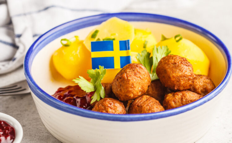 Bild på svit skål med blå kant innehållande köttbullar, potatis och en svensk flagga.